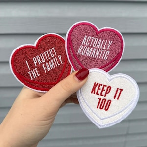 May include: Three heart-shaped patches with embroidered text. One red patch reads "I PROTECT THE FAMILY", a pink patch says "ACTUALLY ROMANTIC", and a white patch says "KEEP IT 100". Each patch has a contrasting border.