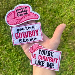 May include: Three embroidered patches with the text "You're a Cowboy Like Me". One patch is shaped like a pink cowboy hat. The other two are rectangular with the text in pink and black. The patches are white with a pink border.