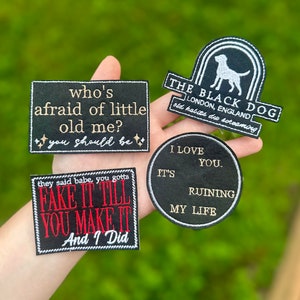 Pode incluir: Uma coleção de patches pretos bordados com costuras brancas. Os patches apresentam frases como "who's afraid of little old me?" e "Fake it till you make it." Um patch tem a silhueta de um cão.