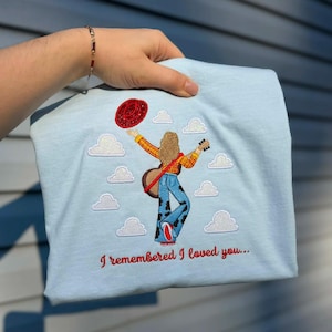 May include: A light blue t-shirt featuring an embroidered design of a person with long hair wearing a yellow plaid shirt and cow-print bell-bottoms, holding a guitar and a red hat, surrounded by white clouds with the text I remembered I loved you.
