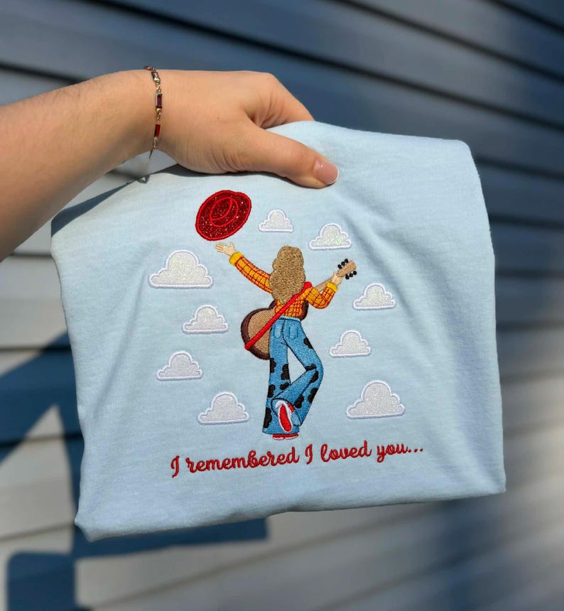 May include: A light blue t-shirt featuring an embroidered design of a person with long hair wearing a yellow plaid shirt and cow-print bell-bottoms, holding a guitar and a red hat, surrounded by white clouds with the text I remembered I loved you.