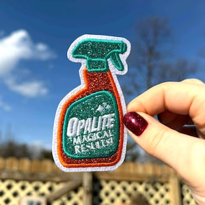 Könnte beinhalten: Ein Aufnäher in Form einer Sprühflasche mit dem weißen Text "OPALITE MAGICAL RESULTS". Der Aufnäher hat einen grünen Glitzeraufsatz, einen roten Glitzerkörper und einen weißen Rand. Vor blauem Himmel gehalten.