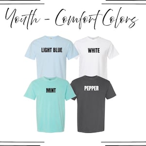 May include: Four short-sleeved t-shirts arranged in a grid, labeled Youth - Comfort Colors. The shirts are light blue, white, mint, and pepper gray. Each shirt features its respective color name printed in bold black or white text across the chest.