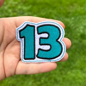 May include: A teal number 13 patch with a black outline and white border. The patch is embroidered with a teal fill and a black outline, set against a white background. The patch is held in a hand.