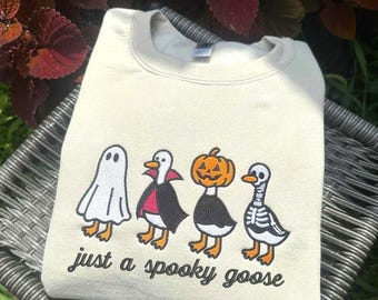 May include: Cream-colored sweatshirt featuring four cartoon geese dressed in Halloween costumes: a ghost, a vampire, a jack-o'-lantern, and a skeleton. The phrase "just a spooky goose" is printed below the geese in black script.