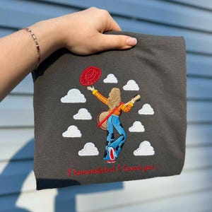 May include: A dark gray t-shirt featuring an embroidered design of a person with long hair, wearing a yellow plaid shirt and blue cow-print bell-bottom pants, holding a guitar and a red hat, surrounded by white clouds with the text I remembered I loved you.