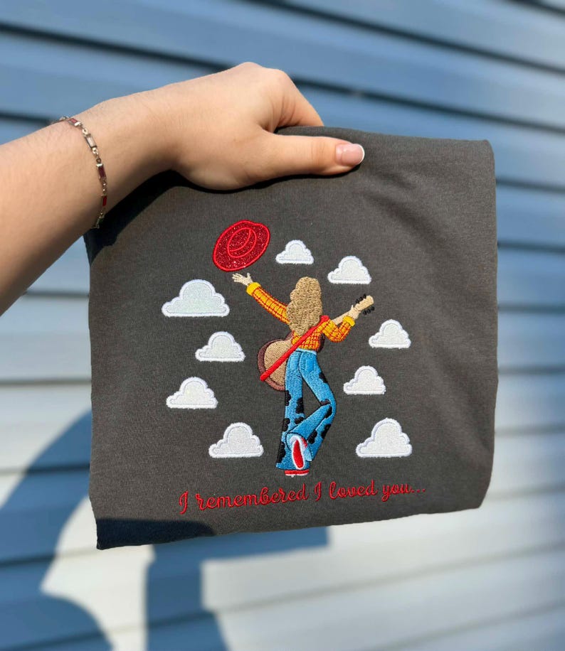 May include: A dark gray t-shirt featuring an embroidered design of a person with long hair, wearing a yellow plaid shirt and blue cow-print bell-bottom pants, holding a guitar and a red hat, surrounded by white clouds with the text I remembered I loved you.