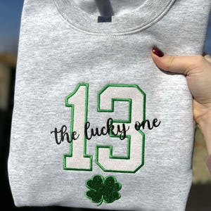 May include: A heather gray sweatshirt with the number 13 in green and white, the words "the lucky one" in black script, and a green shamrock. The sweatshirt is held up by a hand.