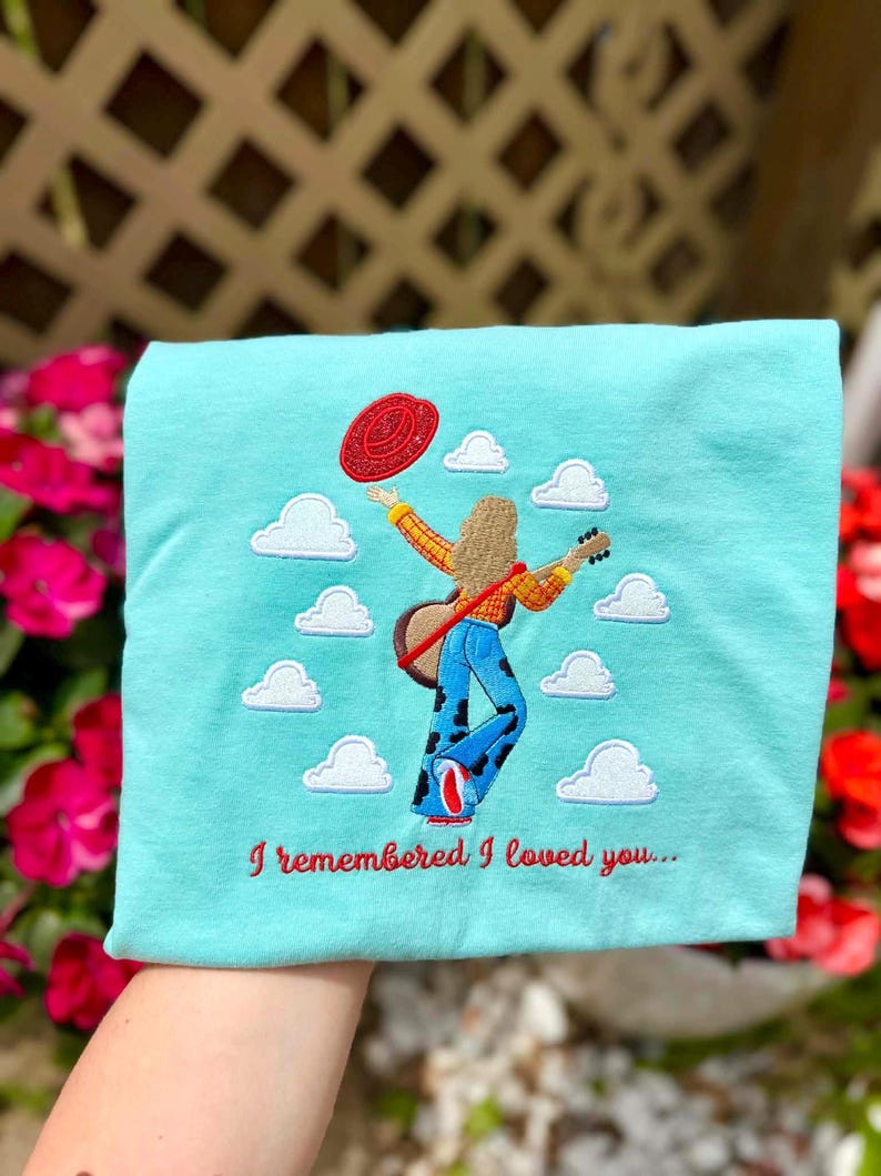 May include: A light blue t-shirt featuring an embroidered design of a person with long hair wearing a yellow plaid shirt and cow-print pants, holding a guitar and a red hat, surrounded by white clouds with the text I remembered I loved you...