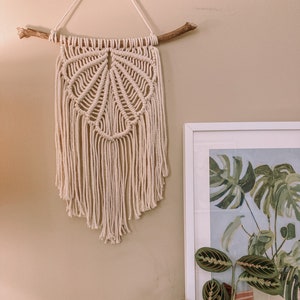 May include: A beige macrame wall hanging with a fan-shaped design, hanging from a branch, with a green plant and a framed print in the background.