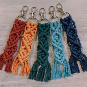 May include: Five colourful macrame keychains with silver key rings. The keychains are in shades of red, orange, green, blue, and navy blue.