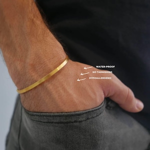 Puede incluir: Una pulsera rígida de metal dorado liso con un diseño minimalista. La imagen incluye texto en blanco y flechas que señalan la pulsera, etiquetándola como WATER-PROOF, NO TARNISHING y HYPOALLERGENIC, destacando sus características principales.