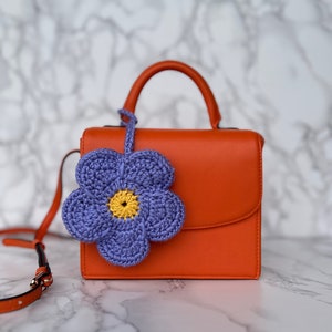 May include: A bright orange leather handbag with a top handle and a detachable shoulder strap. A purple crocheted flower with a yellow center is attached to the bag.
