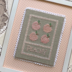 Może przedstawiać: Ramka z haftem krzyżykowym o tematyce brzoskwiniowej. Haft przedstawia cztery różowe brzoskwinie z zielonymi liśćmi i napis "Peachy" w ozdobnym, pochylonym piśmie.