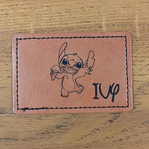 May include: Brown leather patch with a black stitched border. The patch features a cartoon image of Stitch from Lilo & Stitch holding an ice cream cone. The text "I <3 U" is printed below Stitch.