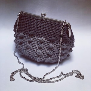 Puede incluir: Un bolso de crochet gris con cierre de plata y correa de cadena. El bolso tiene un diseño texturizado y abultado.