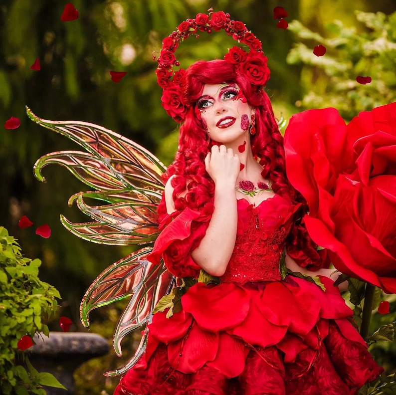 May include: A woman dressed as a fairy with red hair and a red rose crown. She is wearing a red dress with a large red rose attached to the back. She has a pair of large, iridescent fairy wings.