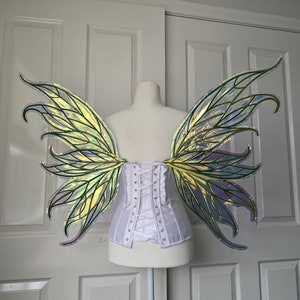 May include: A pair of iridescent fairy wings with a green and blue color scheme. The wings are attached to a white corset.