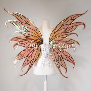 May include: A pair of large, iridescent fairy wings with a peach and gold color scheme. The wings are made of a translucent material and have a delicate, intricate design. The wings are attached to a white corset.