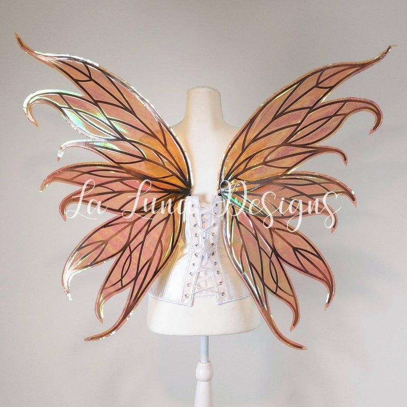 May include: A pair of large, iridescent fairy wings with a peach and gold color scheme. The wings are made of a translucent material and have a delicate, intricate design. The wings are attached to a white corset.