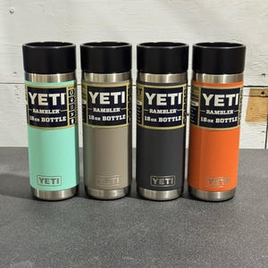 May include: Four YETI Rambler 18 oz bottles in various colors: seafoam green, tan, charcoal gray, and orange. Each bottle has a black lid and the YETI logo. The text "RAMBLER 18 oz BOTTLE" is printed on each bottle.