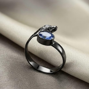 Könnte beinhalten: Ein schwarzer Metallring mit Schlangenmotiv. Der Schlangenkopf umschließt einen blauen, ovalen Edelstein. Der Ring wird vor einem neutralen Stoffhintergrund präsentiert, der die Details des Schmucks hervorhebt. Das Design des Rings ist elegant und einzigartig.