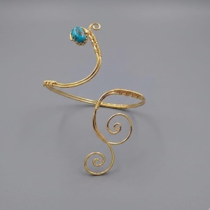May include: A gold-toned wire arm cuff featuring a spiral design and a single oval turquoise-colored stone at one end. The metal wire is wrapped around itself to form decorative swirls and curves, creating an open, adjustable structure.