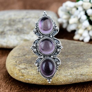Può includere: Un anello argentato con tre pietre preziose ovali viola. L'anello presenta un design a filigrana ornato attorno alle pietre e lungo il gambo. Le pietre preziose sono disposte verticalmente, creando un gioiello di grande effetto.