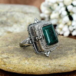 May include: A silver ring with a hinged compartment and a large, emerald green gemstone. The ring is ornate and detailed, with a textured finish.