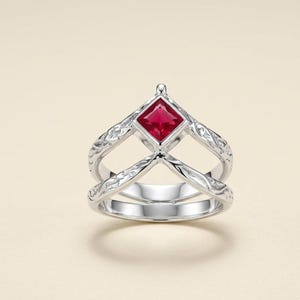 May include: A silver ring with a square, red gemstone at the center. The ring has an intricate, swirling design on the band and around the gemstone. The ring is set against a cream-colored background.