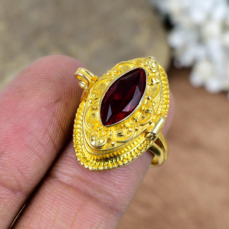 May include: A gold-colored ring with an oval, deep red gemstone. The ring has an ornate, detailed design around the gemstone and on the band. The ring is held by a person's hand.