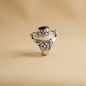 May include: A silver locket ring featuring a smooth, oval black stone set into the top. The band and setting are decorated with intricate metal beadwork and scroll patterns, creating a textured appearance around the central black gemstone.