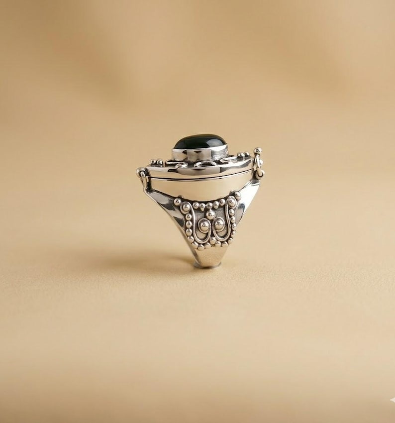 May include: A silver locket ring featuring a smooth, oval black stone set into the top. The band and setting are decorated with intricate metal beadwork and scroll patterns, creating a textured appearance around the central black gemstone.