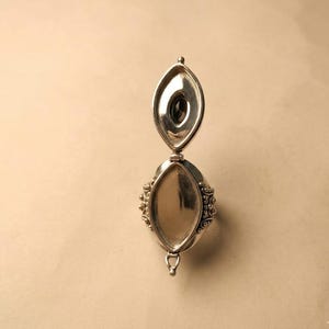 May include: A silver locket ring featuring an elongated marquise-shaped design with a hinged lid that opens vertically. The exterior surface displays intricate beaded metalwork along the sides, while the interior reveals a dark oval stone.