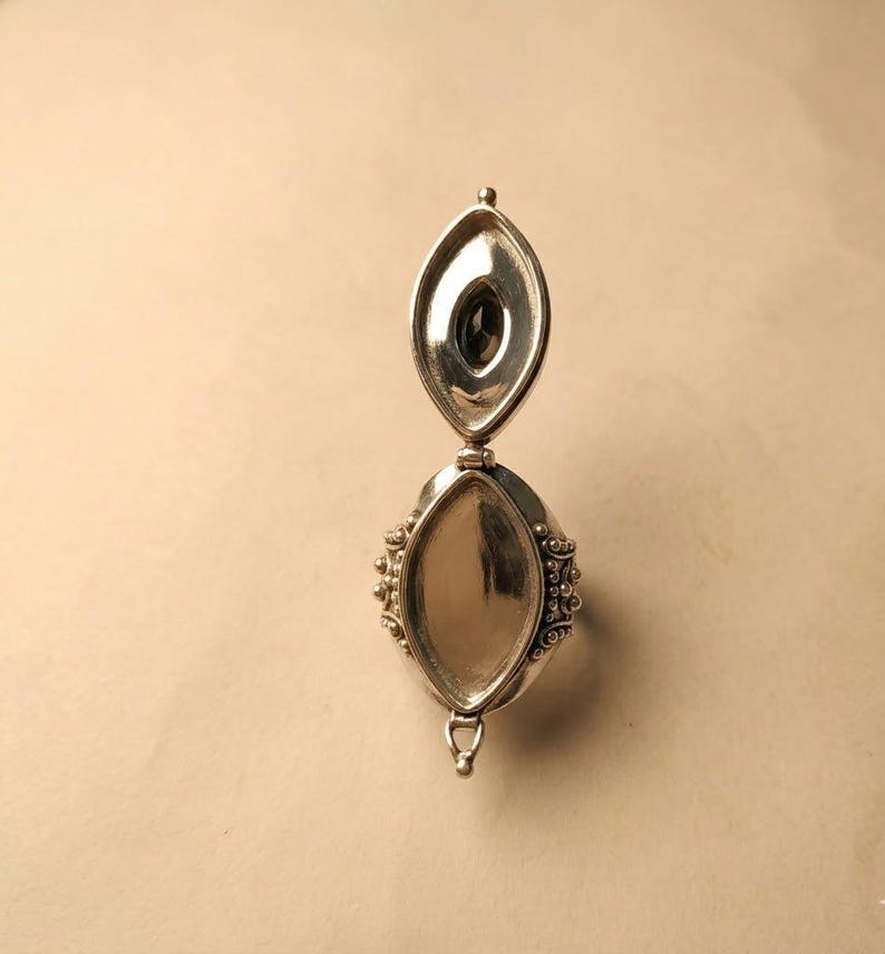 May include: A silver locket ring featuring an elongated marquise-shaped design with a hinged lid that opens vertically. The exterior surface displays intricate beaded metalwork along the sides, while the interior reveals a dark oval stone.