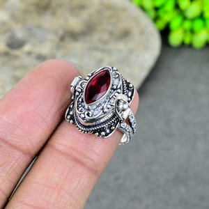 Könnte beinhalten: Ein silberner Ring mit einem verzierten Design und einem roten Edelstein. Der Ring ist ein Ring mit einem Geheimfach, mit einem Scharnierdeckel, der sich öffnet, um einen kleinen Raum im Inneren zu enthüllen.