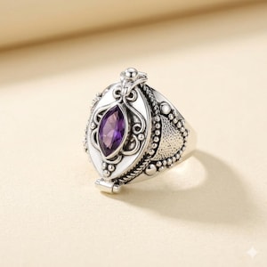 Puede incluir: Anillo plateado con una gema púrpura ovalada. El anillo tiene un diseño detallado con un compartimento con bisagras. La banda tiene un patrón texturizado y el engaste es blanco.