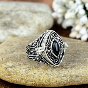 May include: A silver ring with a black stone set in a detailed, ornate design. The ring has a hinged lid that opens to reveal a small compartment.