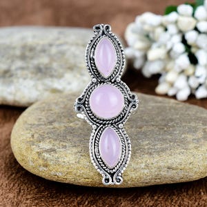 May include: A silver ring with three pale pink gemstones. The ring has an elaborate design with oval and round stones set in a detailed, textured setting. The ring is displayed on a natural stone surface.
