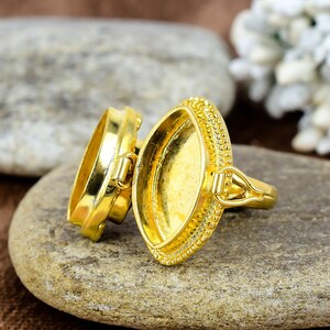 May include: A gold-colored ring with an oval setting and a decorative beaded edge. The ring is open, revealing the interior. The ring is resting on a gray stone surface.