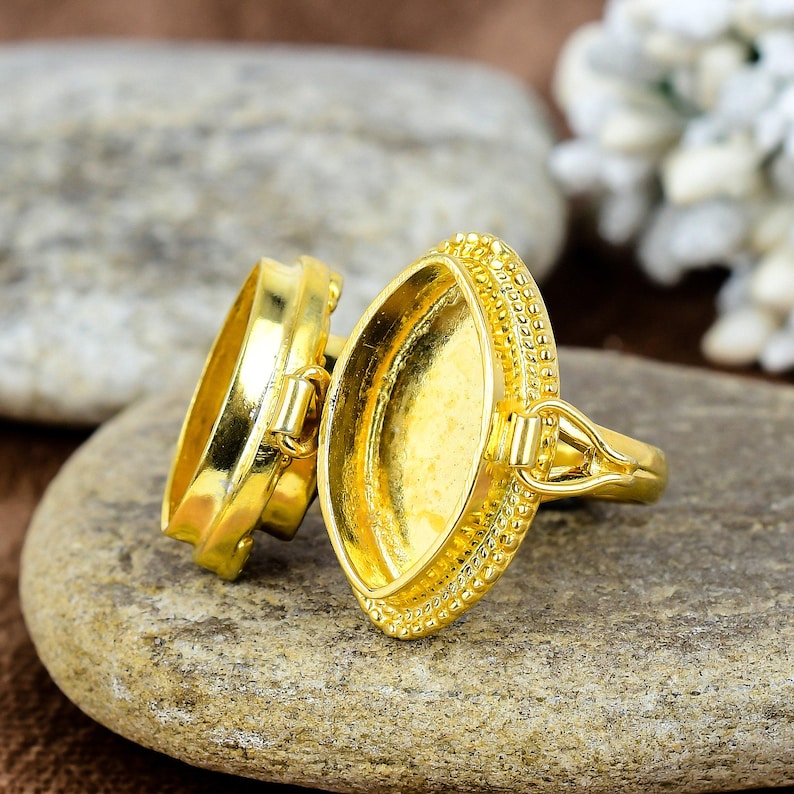 May include: A gold-colored ring with an oval setting and a decorative beaded edge. The ring is open, revealing the interior. The ring is resting on a gray stone surface.
