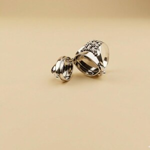 May include: A silver-toned metal ring featuring a hinged locket design that opens to reveal a hollow interior. The exterior surface of the ring is decorated with intricate beaded detailing and small metallic spheres along the top band.