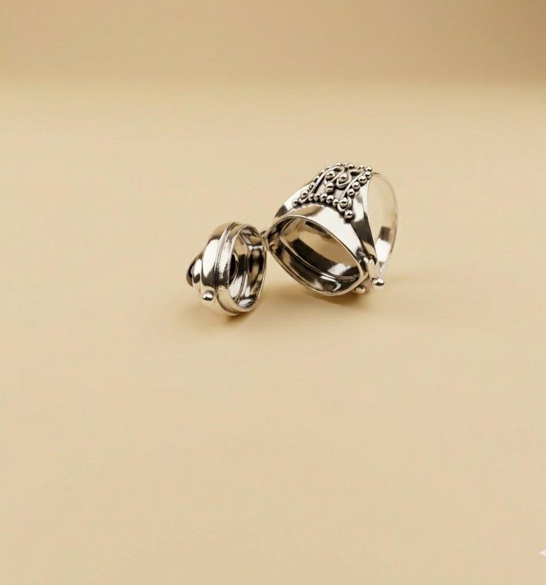 May include: A silver-toned metal ring featuring a hinged locket design that opens to reveal a hollow interior. The exterior surface of the ring is decorated with intricate beaded detailing and small metallic spheres along the top band.