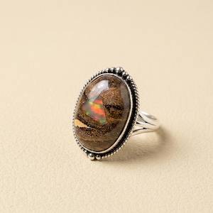 Puede incluir: Anillo de ópalo ovalado con una banda de plata. El ópalo presenta colores marrón, rojo y verde. El anillo tiene un borde con cuentas. El anillo está sobre un fondo neutro.