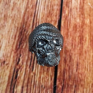 Puede incluir: Un anillo de calavera plateado con detalles intrincados, incluyendo un gorro texturizado y una barba detallada. El anillo se presenta sobre un fondo de madera, resaltando el tema gótico.