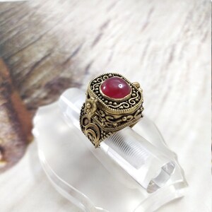 Può includere: Un anello d'oro con una pietra rossa e dettagli intricati. L'anello è una scatola con cerniera e una piccola apertura sul lato.