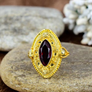 May include: A gold-colored ring with an oval, dark red gemstone. The ring has an ornate, filigree design around the gemstone and on the band. The ring is set on a light brown stone surface.