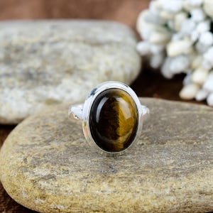 Könnte beinhalten: Ein silberner Ring mit einem ovalen Tigeraugen-Edelstein. Der Edelstein zeigt Braun- und Goldtöne. Der Ring liegt auf einer hellen Steinoberfläche mit unscharfem Hintergrund.