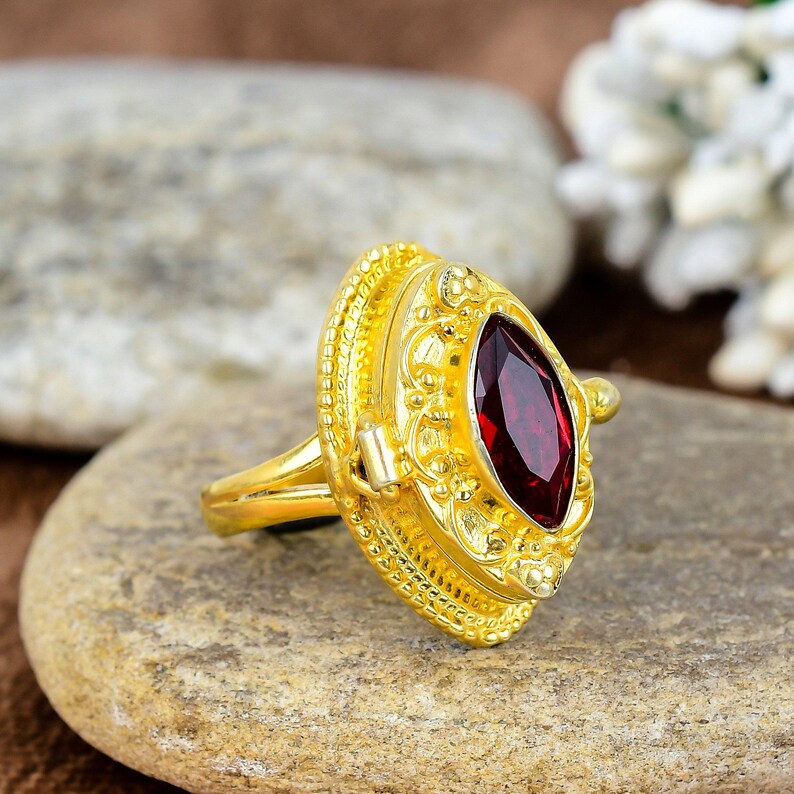 May include: A gold-colored ring with an oval, deep red gemstone. The ring has an ornate, detailed design around the gemstone and on the band. The ring is set on a light brown stone surface.