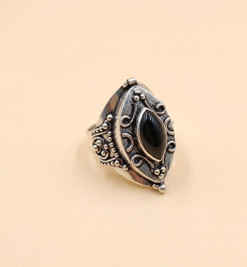 May include: A silver ring featuring an elongated marquise-shaped black stone set in the center. The metal band is decorated with intricate scrollwork, swirling patterns, and small raised bead accents, creating a textured and detailed surface.